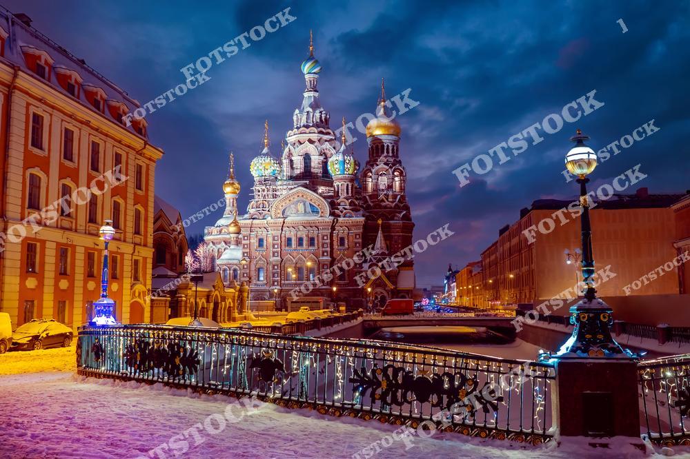 На фотографии изображен Храм Спаса на Крови в зимнюю ночь. Яркая подсветка подчеркивает разноцветные купола и изысканную архитектуру. Заснеженные улицы и мост через канал Грибоедова создают атмосферу зимнего волшебства. Этот храм является одной из главных достопримечательностей Санкт-Петербурга и привлекает множество туристов в любое время года.